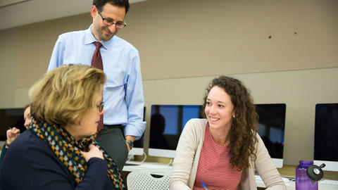 three people enganging with each other, two are sitting at a table with computers in the background, while the third is standing seemingly reviewing the work of the other two.
