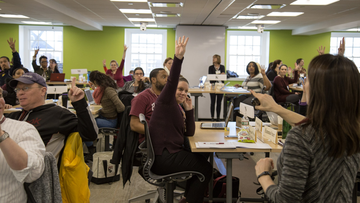 Students in classroom with hands raised and a teacher at the front of the class pointing to a student